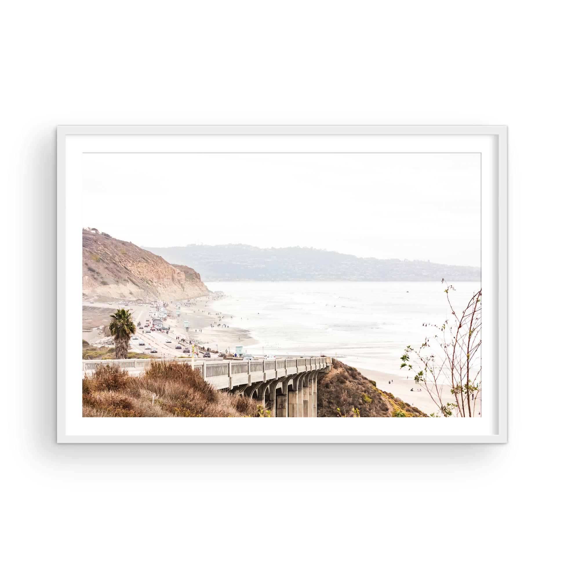 Captivating San Diego coast scene, presented as horizontal beach art. This piece of travel photography features a prominent bridge extending over a wide sandy beach with calm ocean waves. A coastal road winds along the base of rugged cliffs, with a lone coastal palm tree adding a touch of tropical essence to this Southern California seascape print. Photograph in minimalist mock up