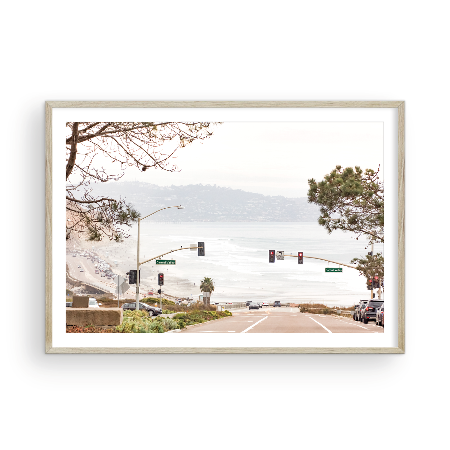 A hazy eye-level shot of San Diego Streets looks down a sloping road featuring red traffic lights and "Carmel Valley" signs, capturing a Local Surf Print vibe. The foreground of this California Wall Art showcases landscape and cars, framed by tree branches at the top, adding to the San Diego Photo aesthetic. The lighting suggests a daytime, perfect for Minimalist Beach Wall Art. This horizontal San Diego print makes a great piece of horizontal beach wall decor. Photograph in minimalist mock up