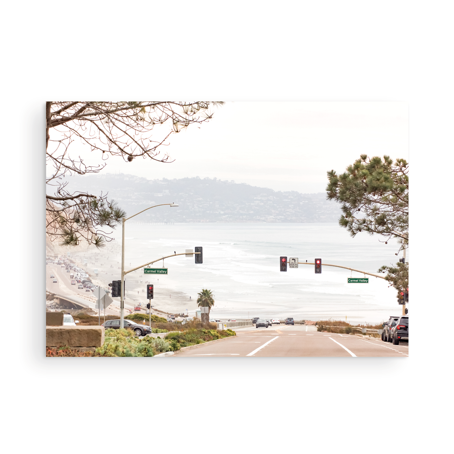 A hazy eye-level shot of San Diego Streets looks down a sloping road featuring red traffic lights and "Carmel Valley" signs, capturing a Local Surf Print vibe. The foreground of this California Wall Art showcases landscape and cars, framed by tree branches at the top, adding to the San Diego Photo aesthetic. The lighting suggests a daytime, perfect for Minimalist Beach Wall Art. This horizontal San Diego print makes a great piece of horizontal beach wall decor. Photograph in minimalist mock up