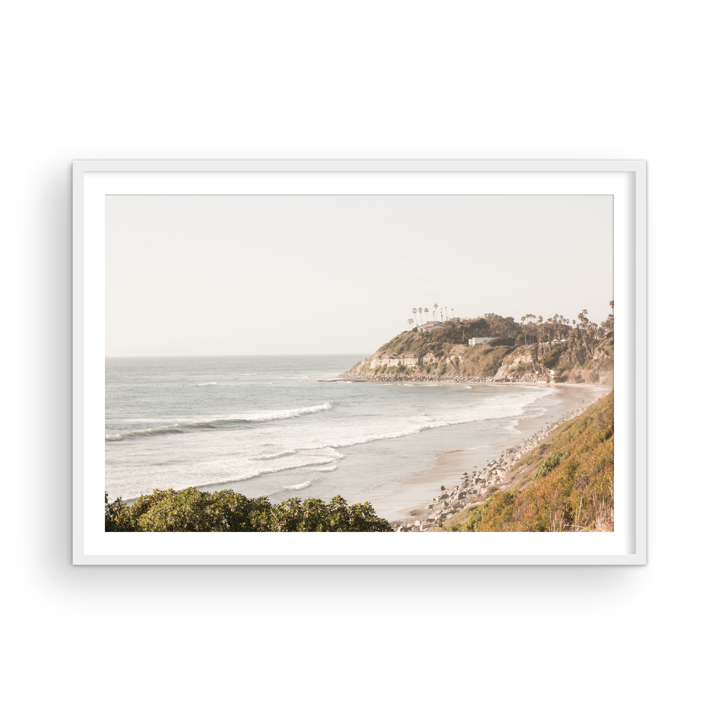 Fine art of Swamis Beach, San Diego California horizontal print of hazy high-angle landscape beach photography captures a light blue ocean meeting a sandy beach with many beach goers. To the right, tall tan and brown cliffs rise as iconic coastal scenery. Photograph in minimalist framed mock up.