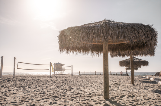 Landscape coastal photography capturing a thatched beach umbrella on a sandy beach, volleyball nets and a lifeguard tower in the distance, with ocean views on the horizon. Photograph in minimalist mock up.