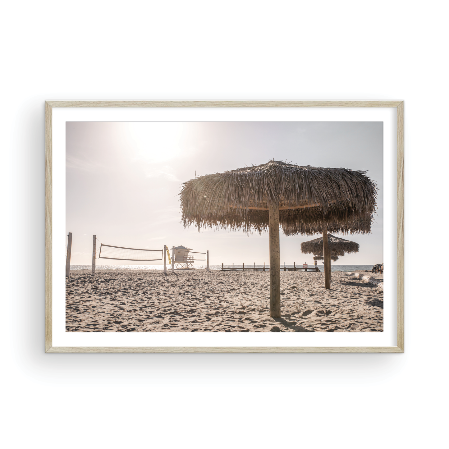 Landscape coastal photography capturing a thatched beach umbrella on a sandy beach, volleyball nets and a lifeguard tower in the distance, with ocean views on the horizon. Photograph in minimalist mock up.