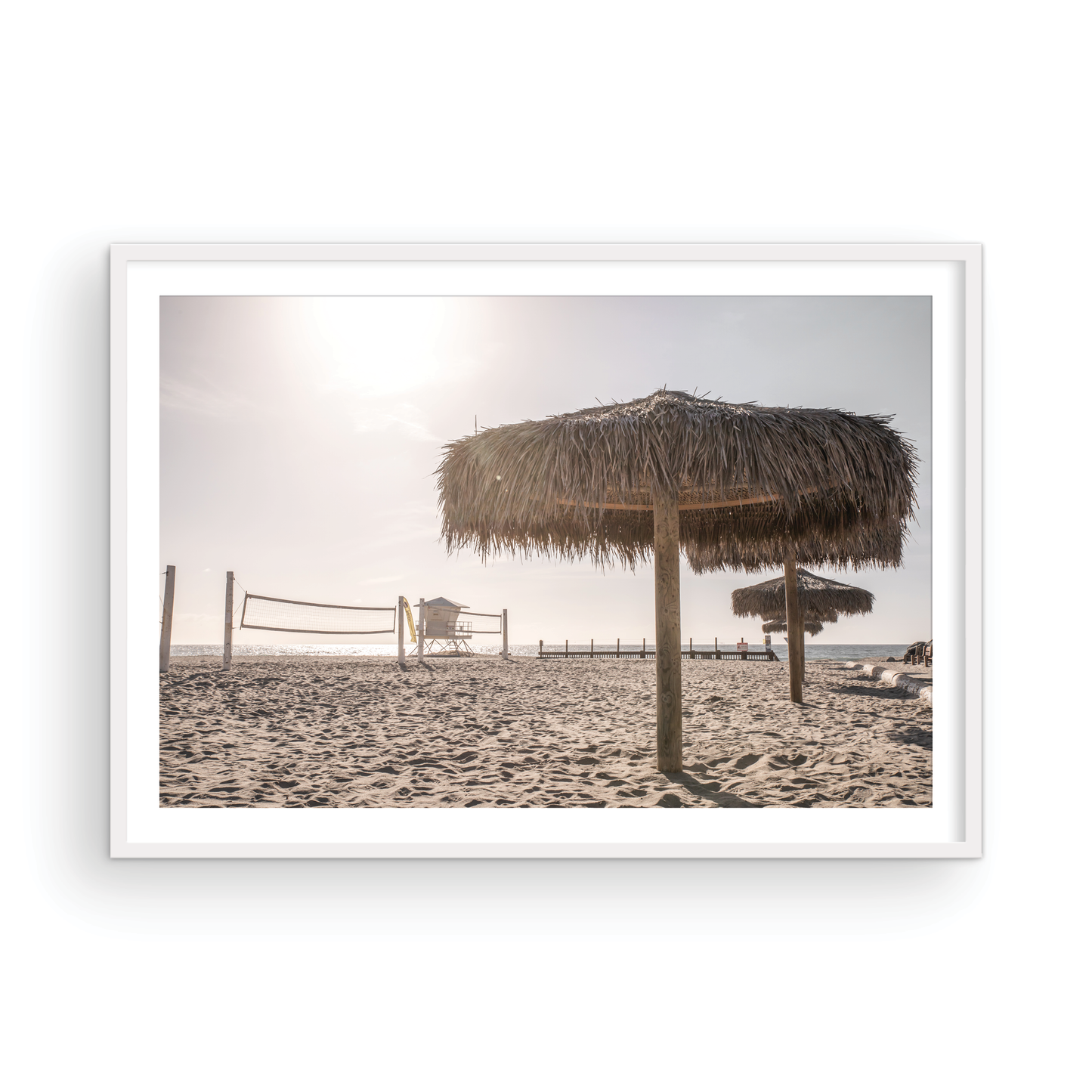 Landscape coastal photography capturing a thatched beach umbrella on a sandy beach, volleyball nets and a lifeguard tower in the distance, with ocean views on the horizon. Photograph in minimalist mock up.