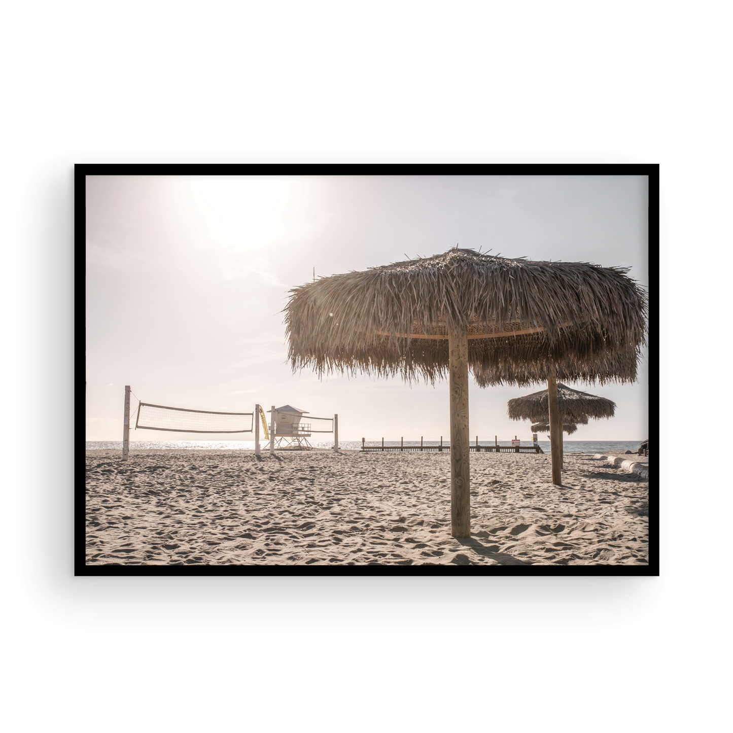 Landscape coastal photography capturing a thatched beach umbrella on a sandy beach, volleyball nets and a lifeguard tower in the distance, with ocean views on the horizon. Photograph in minimalist mock up.