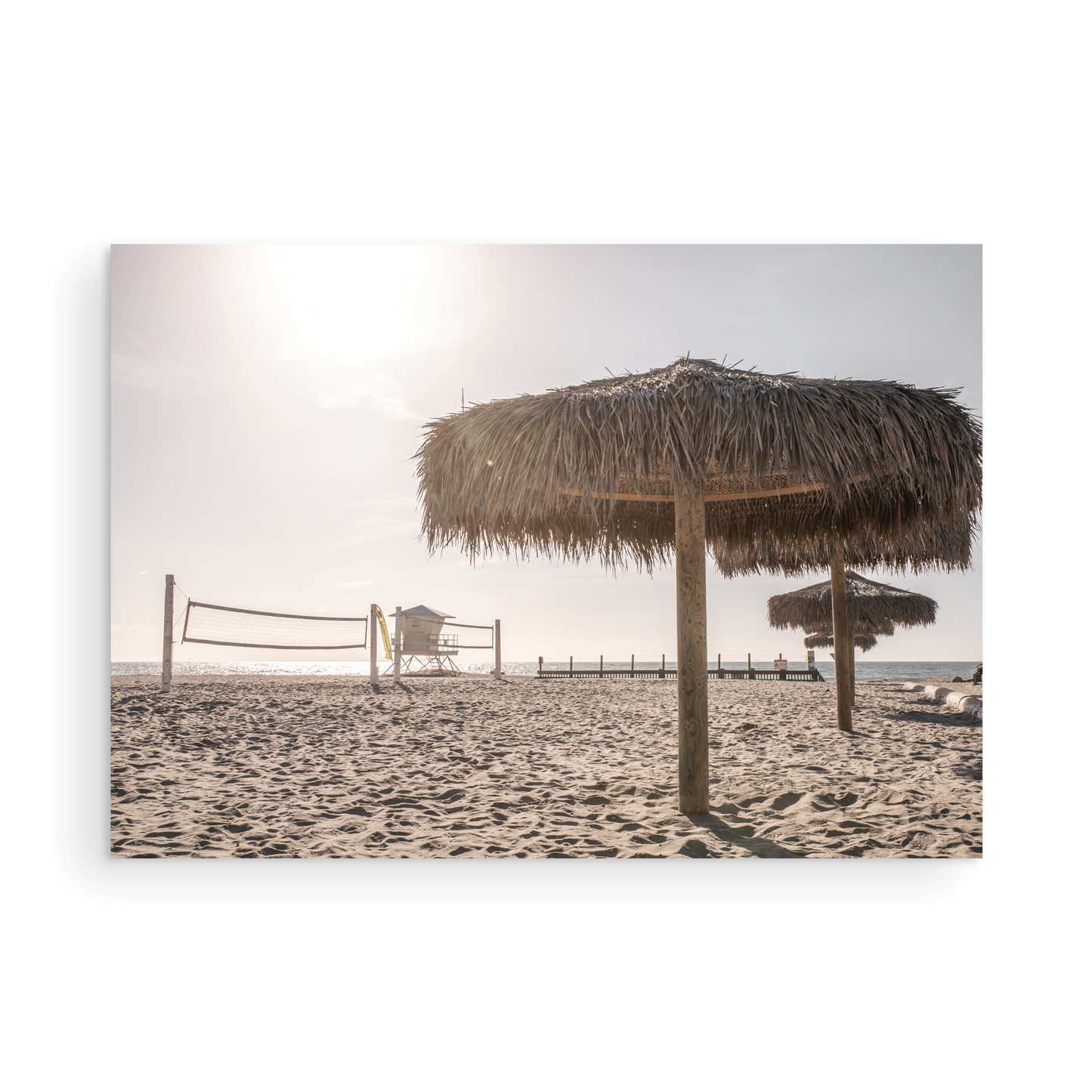 Landscape coastal photography capturing a thatched beach umbrella on a sandy beach, volleyball nets and a lifeguard tower in the distance, with ocean views on the horizon. Photograph in minimalist mock up.