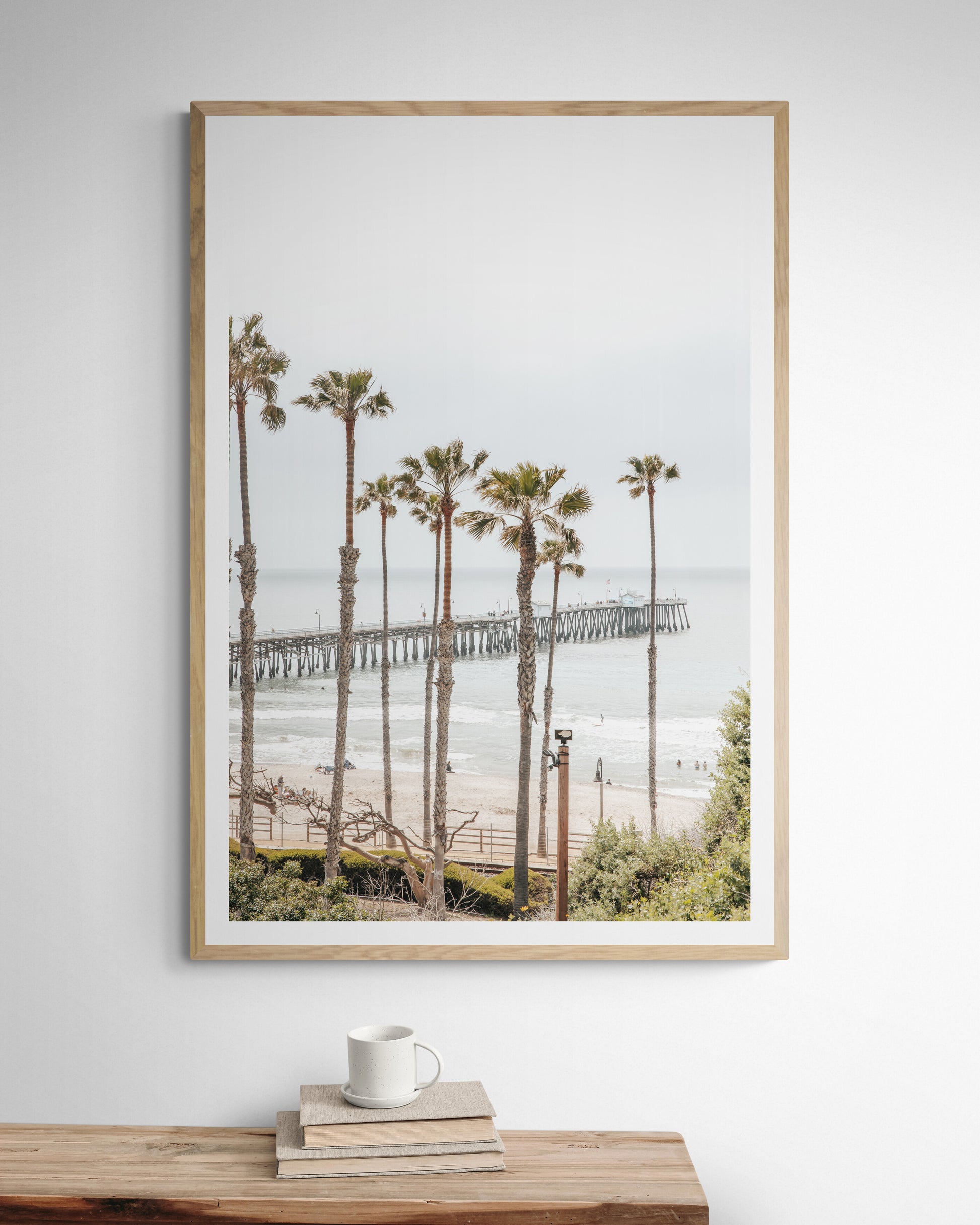 A vertical landscape coastal photography shot of the San Clemente Pier from behind a lush line of tall palm trees. This beachscape print highlights the California coast and is ideal for large beach art or a beach theme wall decor, with subtle ocean waves. Photograph in minimalist mock up.