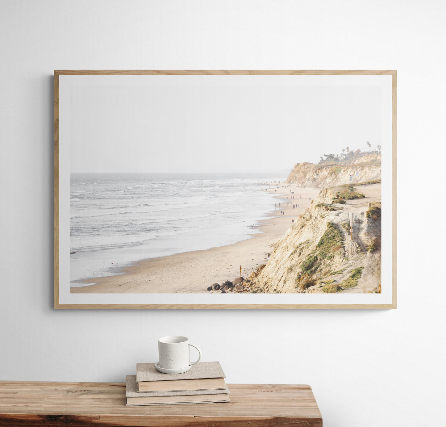 Fine art of Torrey Pines, San Diego California horizontal print of hazy high-angle landscape beach photography captures a light blue ocean meeting a sandy beach with many beach goers. To the right, tall tan and brown cliffs rise as iconic coastal scenery. Photograph in minimalist framed mock up.