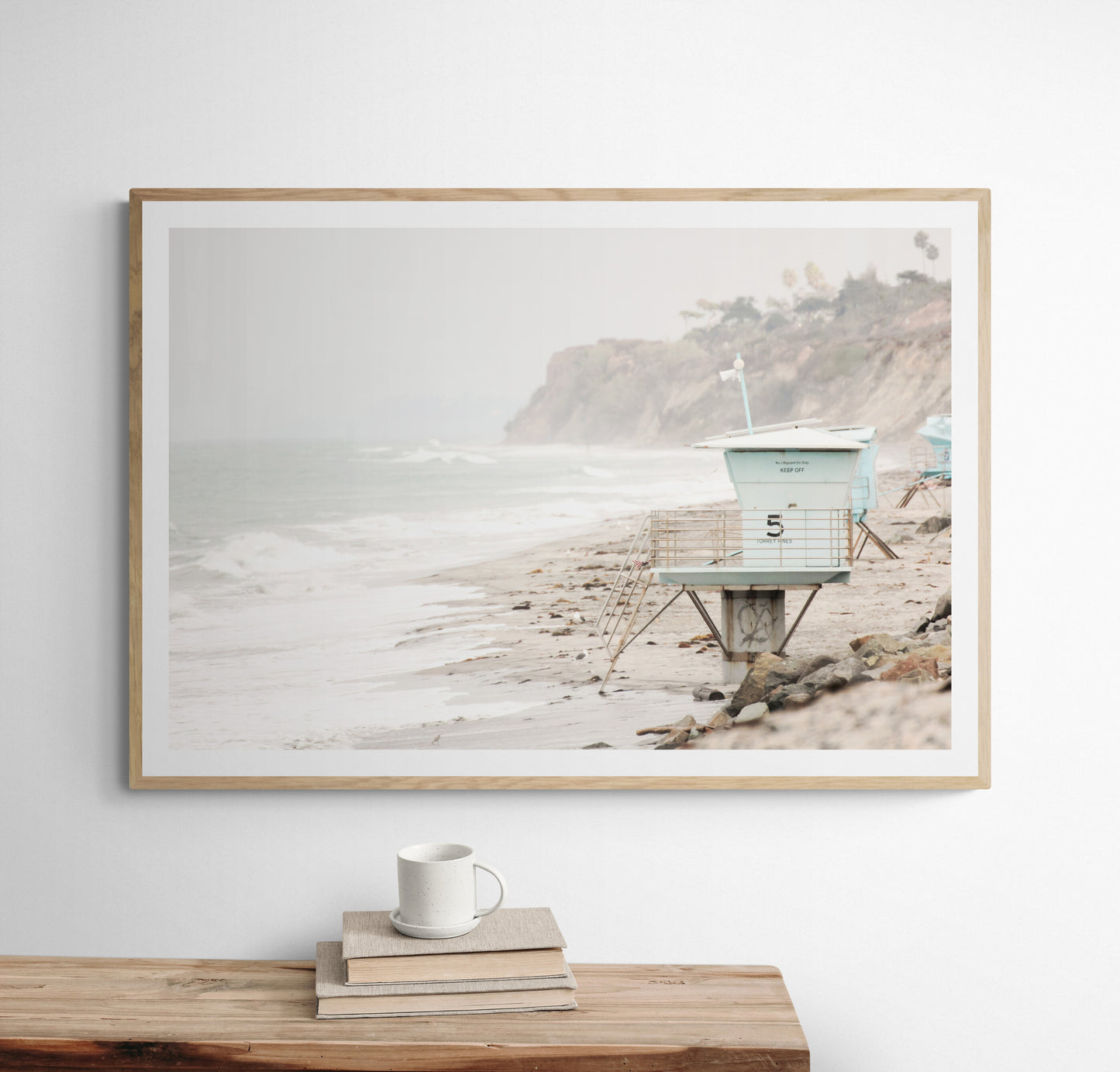 A serene piece of beach photography showcasing a light blue lifeguard tower, number 5, on the San Diego coast. This pastel beach art captures the iconic tower standing on wet sand with gentle ocean waves receding, under a hazy, overcast sky with distant cliffs. A perfect landscape lifeguard tower print for coastal theme wall art. Photograph in minimalist surf art mock up.