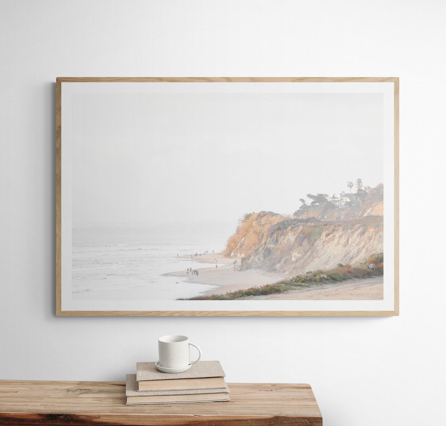 Fine art of Torrey Pines, San Diego California horizontal print of hazy high-angle landscape beach photography captures a light blue ocean meeting a sandy beach with many beach goers. To the right, tall tan and brown cliffs rise as iconic coastal scenery. Photograph in minimalist framed mock up.