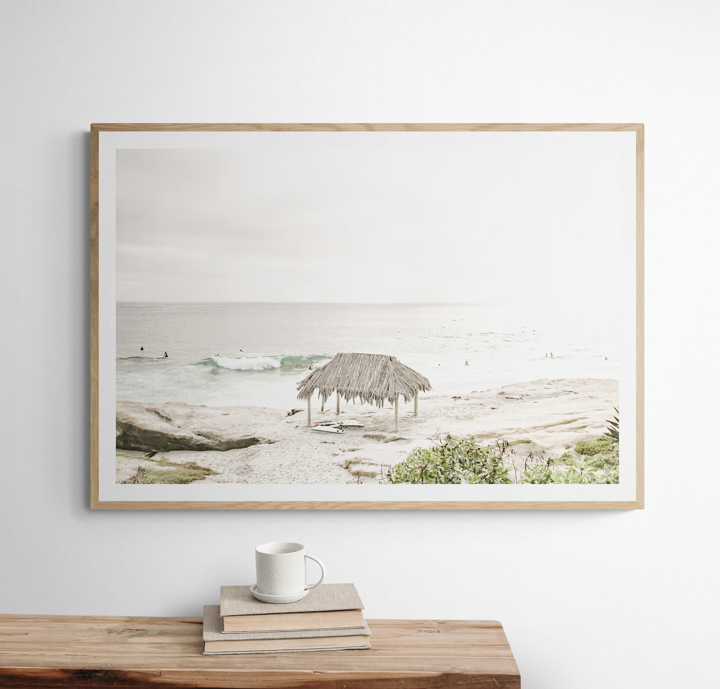 Fine art of WindanSea Beach, La Jolla California horizontal landscape print by Southern California photographer Kim Dybczak of Salt Creek Prints, featuring a bright, slightly desaturated image shows Windansea Beach in La Jolla, California. A thatched beach hut stands on the sandy shore in the left foreground. Further down the beach, a surfer carrying a teal surfboard walks towards the ocean where several other surfers are visible in the water. Photograph in minimalist framed mock up