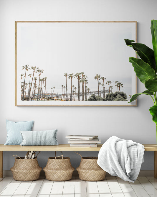Horizontal landscape coastal photography of many palm trees in the foreground, with the San Clemente Pier extending into the ocean in the background. This large landscape beach art conveys strong beach vibes. Photograph in minimalist mock up.