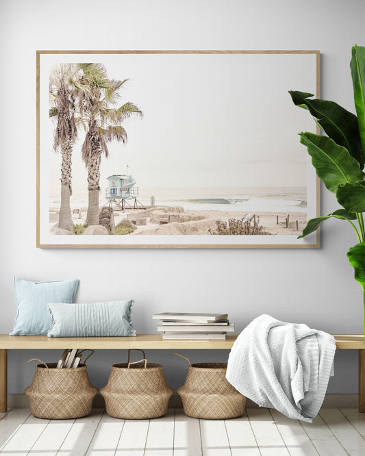 Dreamy coastal palm tree print featuring two tall palm trees with shaggy trunks in the foreground and a pastel blue lifeguard tower in the distance. This horizontal tropical print is captured at Seaside Beach on the Southern California coast, is bathed in bright, hazy sunlight, creating a pastel beach art effect. Evokes serene summer vibes, perfect for beach house decor. Photograph in minimalist mock up.