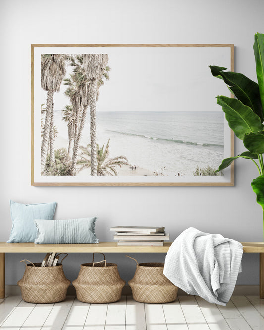 Dreamy coastal palm tree print featuring several tall palm trees with shaggy trunks, and surfers walking down the tranquil beach. This vertical tropical print is captured at Swamis Beach on the Southern California coast, is bathed in bright, hazy sunlight, creating a pastel beach art effect. Evokes serene summer vibes, perfect for beach house decor. Photograph in minimalist surf art mock up.
