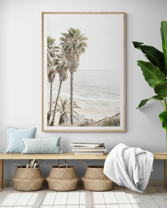 Dreamy coastal palm tree print featuring several tall palm trees with shaggy trunks, next to a staircase down to a tranquil beach. This vertical tropical print is captured at Swamis Beach on the Southern California coast, is bathed in bright, hazy sunlight, creating a pastel beach art effect. Evokes serene summer vibes, perfect for beach house decor. Photograph in minimalist surf art mock up.