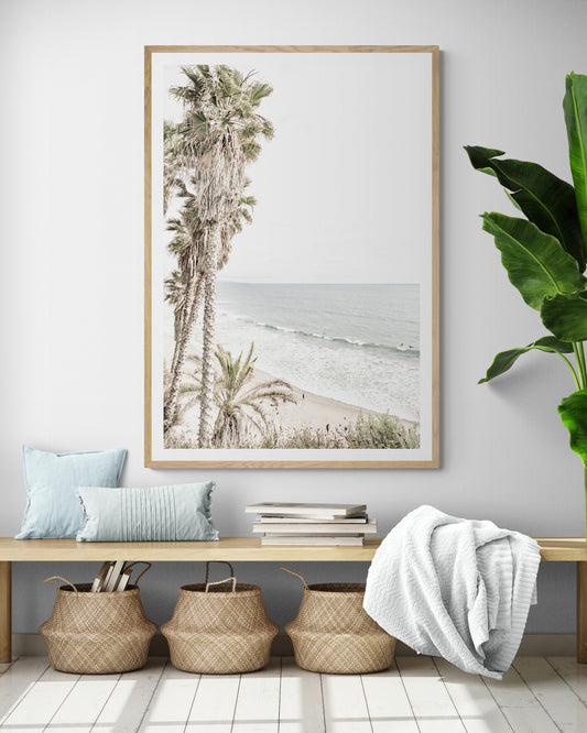 Dreamy coastal palm tree print featuring several tall palm trees with shaggy trunks, lining a gentle slope down to a tranquil beach. This vertical tropical print is captured at Swamis Beach on the Southern California coast, is bathed in bright, hazy sunlight, creating a pastel beach art effect. Evokes serene summer vibes, perfect for beach house decor. Photograph in minimalist surf art mock up.