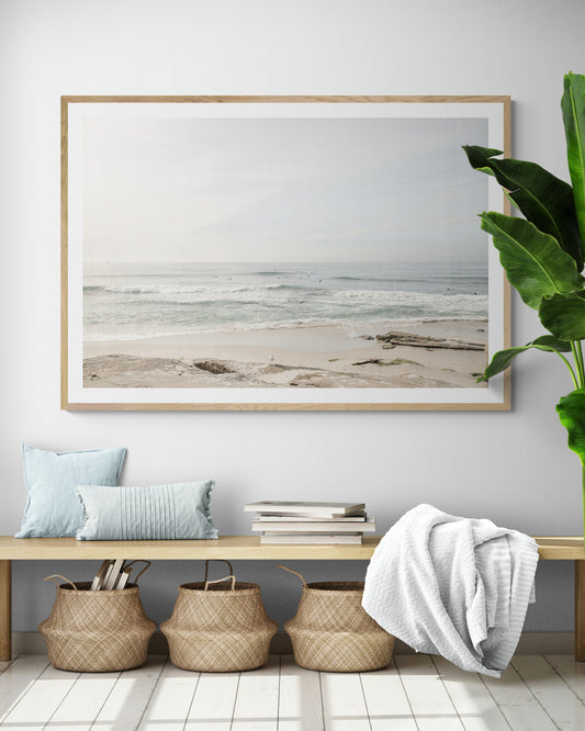 Fine art of horizontal landscape print by Southern California photographer Kim Dybczak of Salt Creek Prints, featuring a bright image captures a pale sandy beach giving way to a vast, light gray ocean with gentle waves. Several surfers are scattered across the water, some sitting on their boards and others riding small waves. The sky is a uniform, pale white, creating a soft, overcast atmosphere, located in Windansea Beach in La Jolla, California. Photograph in framed minimalist mock up