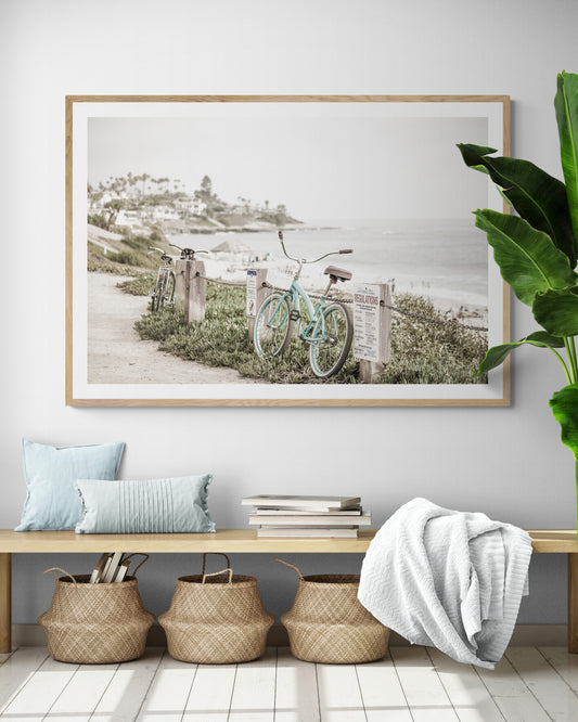 Fine art of horizontal landscape print by Southern California photographer Kim Dybczak of Salt Creek Prints, the image shows a light blue pastel beach cruiser leaning against a wooden post and chain fence. Another bicycle is visible behind it. The beach is in the background with people and a beach hut, located in Windansea Beach in La Jolla, California. Photograph in framed minimalist entry way mock up