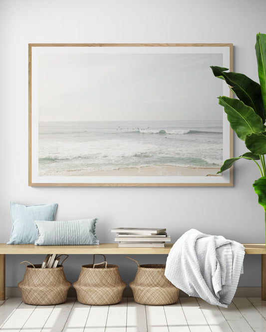 Fine art of horizontal landscape print by Southern California photographer Kim Dybczak of Salt Creek Prints, featuring a bright image captures a pale sandy beach giving way to a vast, light gray ocean with gentle waves. Several surfers are scattered across the water, some sitting on their boards and others riding small waves. The sky is a uniform, pale white, creating a soft, overcast atmosphere, located in Windansea Beach in La Jolla, California. Photograph in framed minimalist entry way mock up