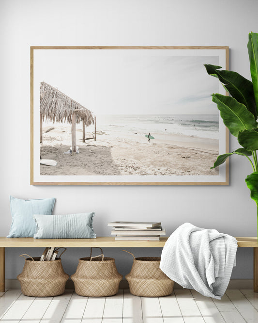 Fine art of WindanSea Beach, La Jolla California horizontal landscape print by Southern California photographer Kim Dybczak of Salt Creek Prints, featuring a bright, slightly desaturated image shows Windansea Beach in La Jolla, California. A thatched beach hut stands on the sandy shore in the left foreground. Further down the beach, a surfer carrying a teal surfboard walks towards the ocean where several other surfers are visible in the water. Photograph in minimalist entry way ,framed mock up