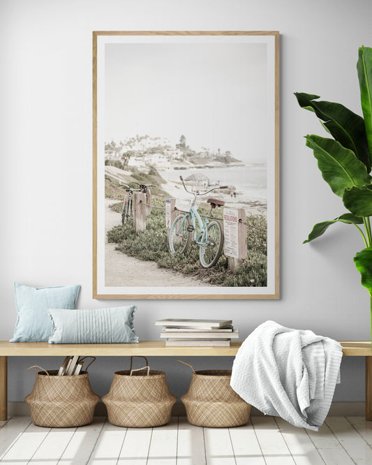 Fine art of horizontal landscape print by Southern California photographer Kim Dybczak of Salt Creek Prints, the image shows a light blue pastel beach cruiser leaning against a wooden post and chain fence. Another bicycle is visible behind it. The beach is in the background with people and a beach hut, located in Windansea Beach in La Jolla, California. Photograph in framed minimalist mock up