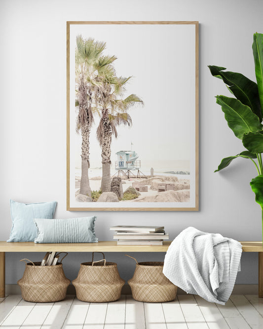 Dreamy coastal palm tree print featuring two tall palm trees with shaggy trunks in the foreground and a pastel blue lifeguard tower centered in the distance. This vertical tropical print is captured at Seaside Beach on the Southern California coast, is bathed in bright, hazy sunlight, creating a pastel beach art effect. Evokes serene summer vibes, perfect for beach house decor. Photograph in minimalist mock up.
