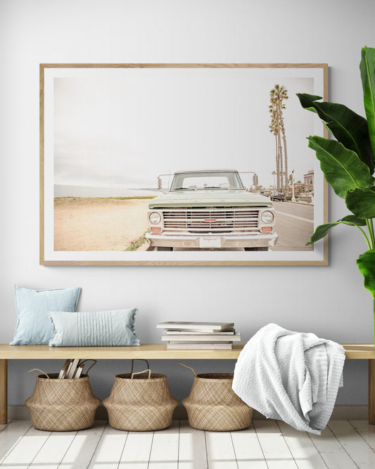 Nostalgic Southern California beach art featuring a classic light green Ford F-Series pickup truck parked by the San Diego coast. This horizontal surf print captures the vintage vehicle with a backdrop of iconic tall palm trees, a hazy beach, and the ocean, embodying surf vibes and perfect for coastal farmhouse decor. Photograph in minimalist framed mock up.