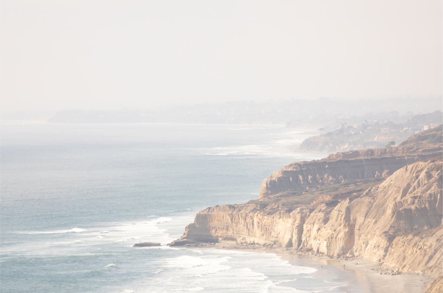 Fine art of Torrey Pines, San Diego California horizontal print of hazy high-angle landscape beach photography captures a light blue ocean meeting a sandy beach with many beach goers. To the right, tall tan and brown cliffs rise as iconic coastal scenery.
