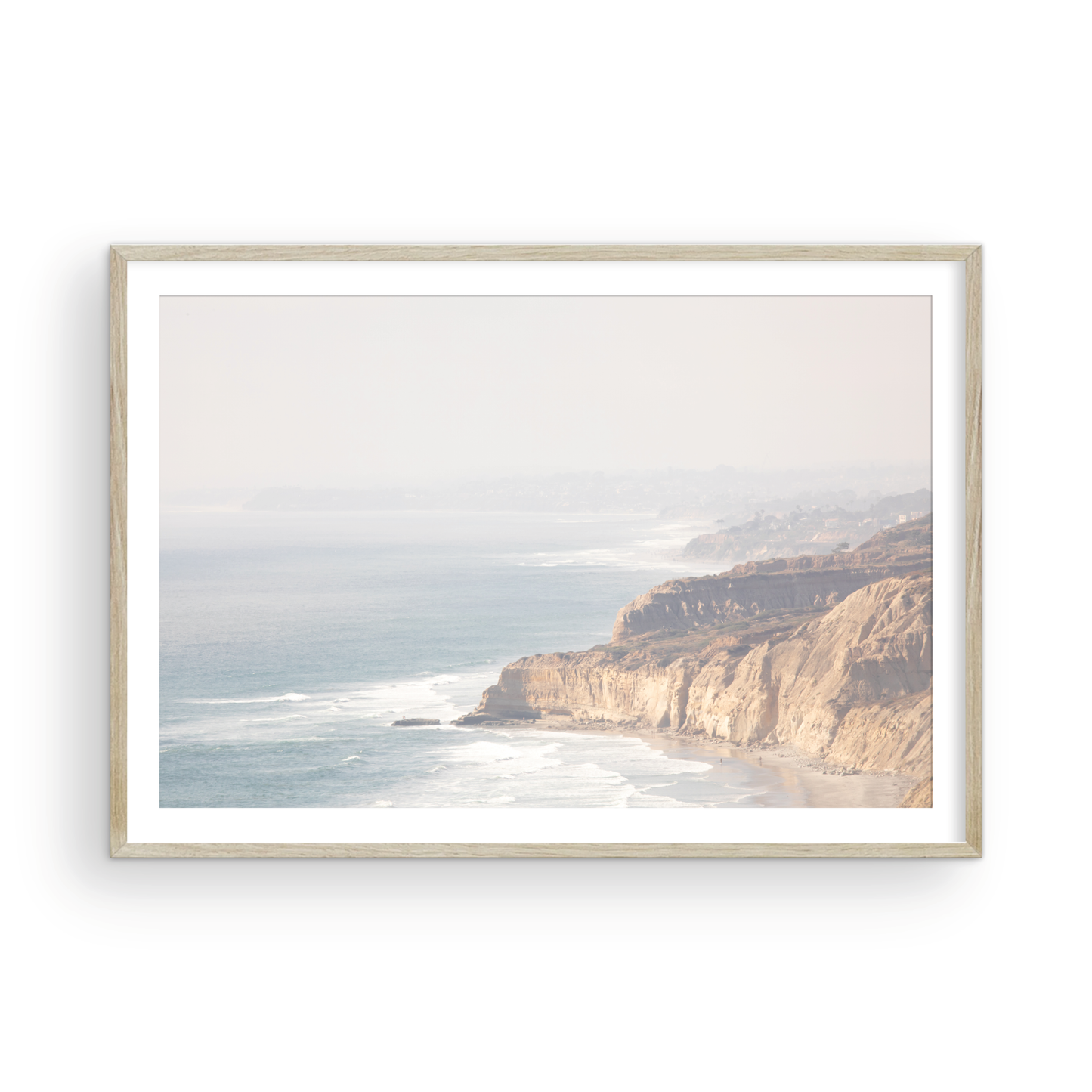 Fine art of Torrey Pines, San Diego California horizontal print of hazy high-angle landscape beach photography captures a light blue ocean meeting a sandy beach with many beach goers. To the right, tall tan and brown cliffs rise as iconic coastal scenery. Photograph in minimalist framed mock up.