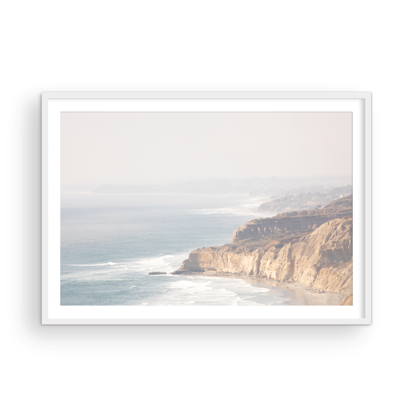 Fine art of Torrey Pines, San Diego California horizontal print of hazy high-angle landscape beach photography captures a light blue ocean meeting a sandy beach with many beach goers. To the right, tall tan and brown cliffs rise as iconic coastal scenery. Photograph in minimalist framed mock up.