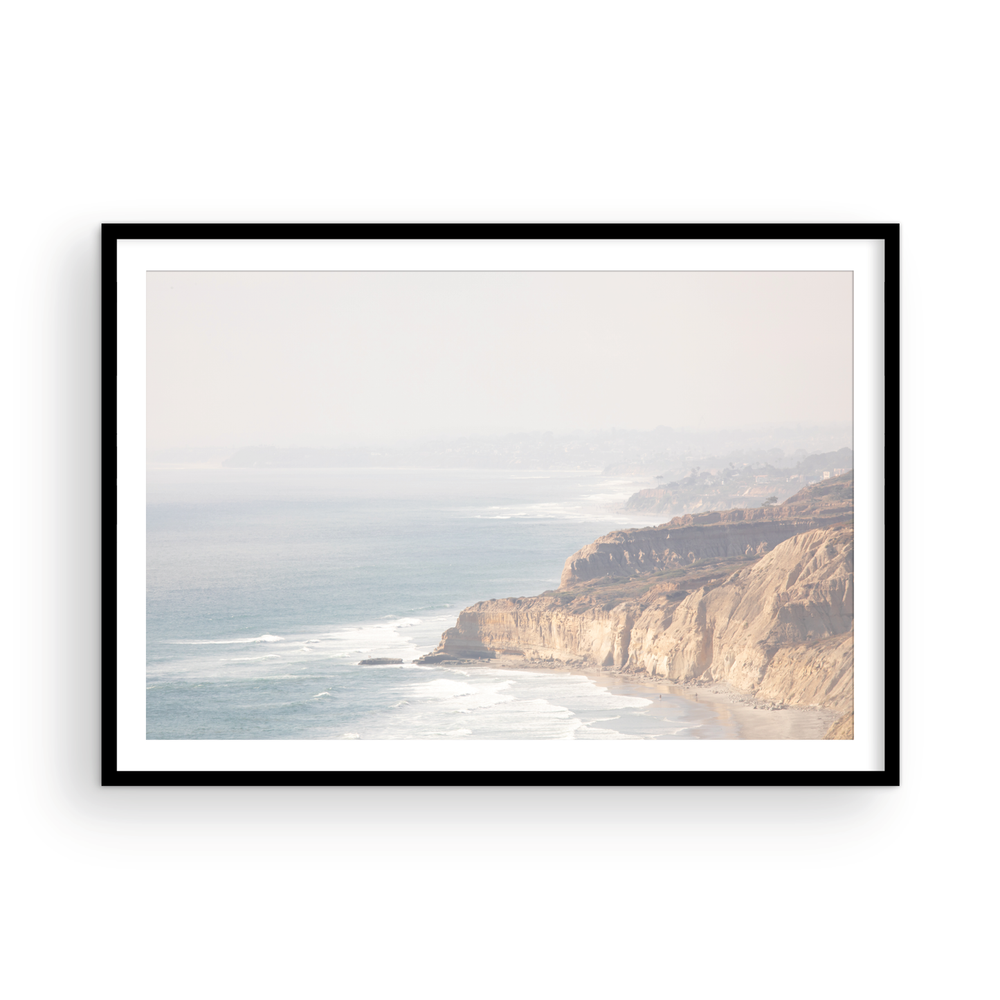 Fine art of Torrey Pines, San Diego California horizontal print of hazy high-angle landscape beach photography captures a light blue ocean meeting a sandy beach with many beach goers. To the right, tall tan and brown cliffs rise as iconic coastal scenery. Photograph in minimalist framed mock up.