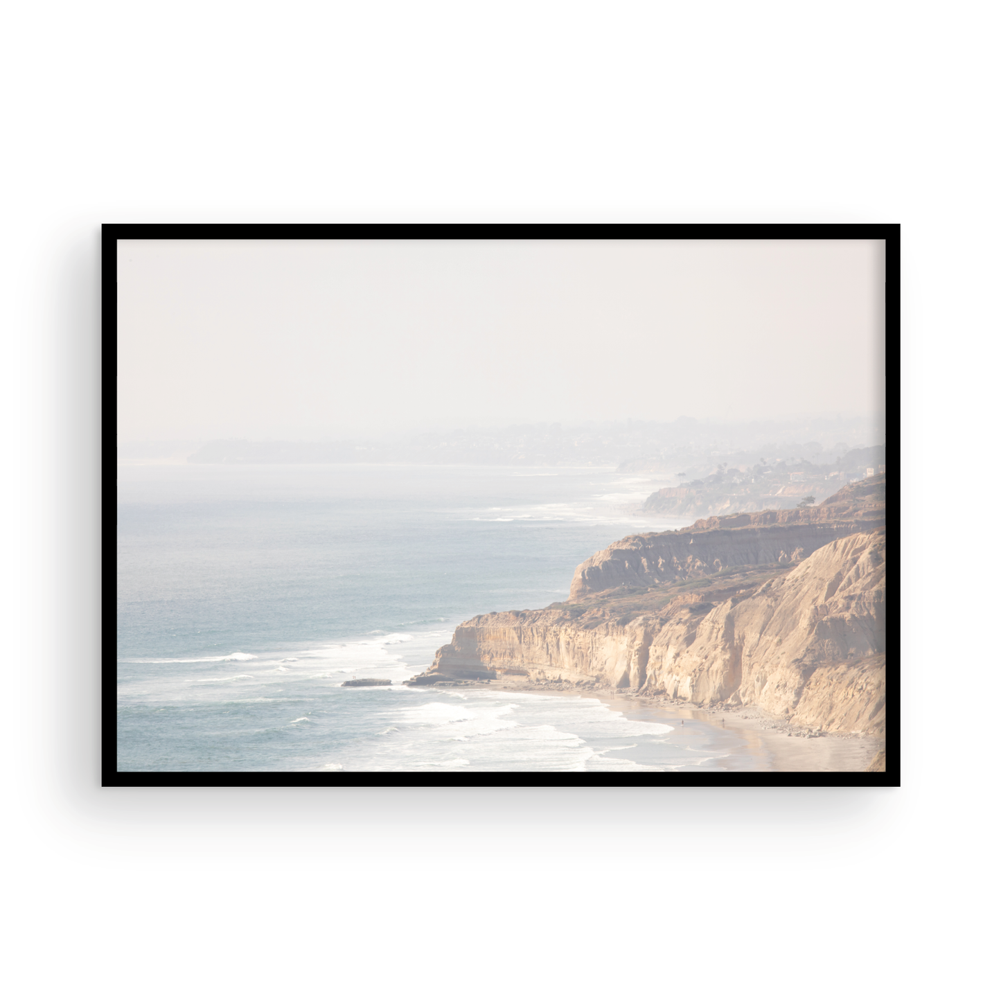 Fine art of Torrey Pines, San Diego California horizontal print of hazy high-angle landscape beach photography captures a light blue ocean meeting a sandy beach with many beach goers. To the right, tall tan and brown cliffs rise as iconic coastal scenery. Photograph in minimalist framed mock up.