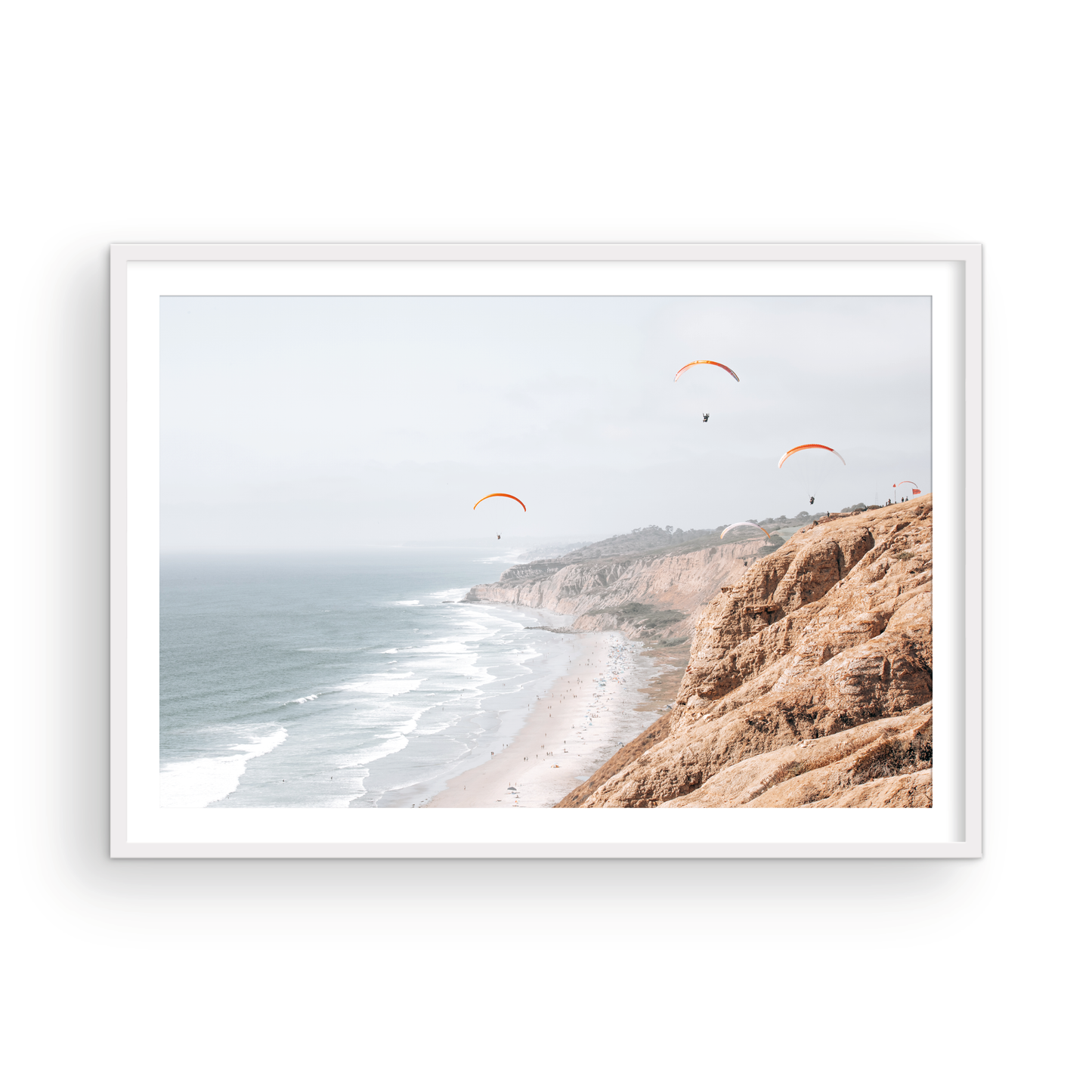 Fine art of Torrey Pines, San Diego California horizontal print of hazy high-angle landscape beach photography captures a light blue ocean meeting a sandy beach with many beach goers. To the right, tall tan and brown cliffs rise as iconic coastal scenery. Four white paragliders gliding in the pale pastel sky, a travel photography moment. The scene emphasizes the cliffs, beach, and the paragliders' presence in the expansive large beach art view. Photograph in minimalist mock up.