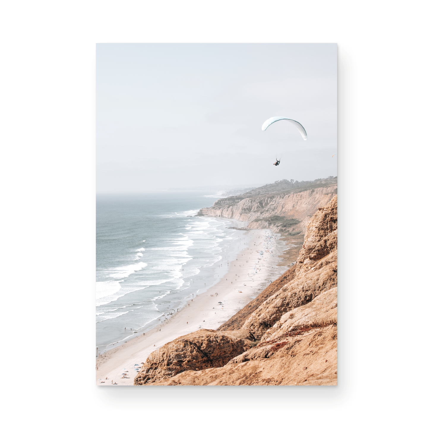 Fine art of Torrey Pines, San Diego California vertical print of hazy high-angle landscape beach photography captures a light blue ocean meeting a sandy beach with many beach goers. To the right, tall tan and brown cliffs rise as iconic coastal scenery. A white paraglider with a pilot flies in the upper right against a pale pastel sky, a travel photography moment. The scene emphasizes the cliffs, beach, and the paraglider's presence in the expansive large beach art view. Photograph in minimalist mock up