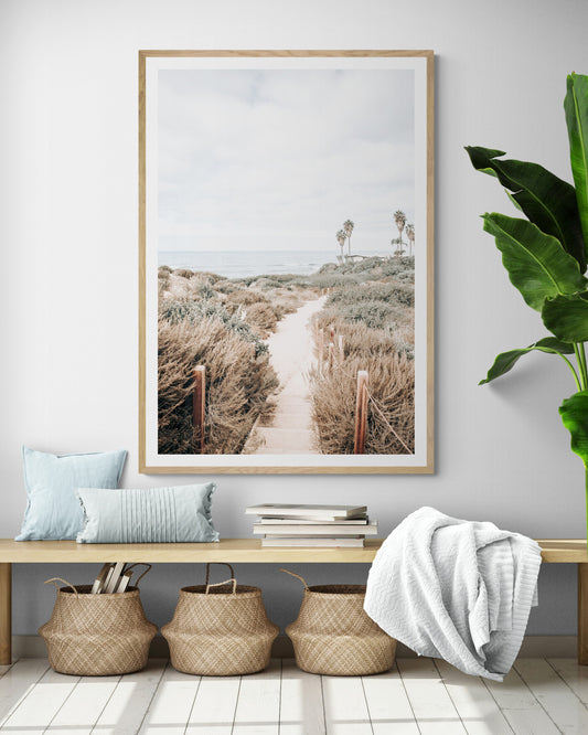 Minimalist vertical beach print showcasing the San Diego Sunset Cliffs, a sandy pathway with rustic wooden steps leading through coastal dune grass towards a calm ocean. This pastel beach art, evocative of the Southern California coastline, features a bright hazy sky and distant palm trees. A tranquil piece of travel photography, ideal for coastal wall art. Photograph in minimalist framed mock up.