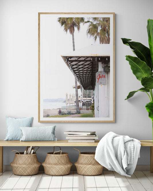 A framed beach scene art print with a wooden structure and palm trees, titled 'Jake's' on the sign, set against a background of a beach and ocean. Photograph in minimalist mock up.