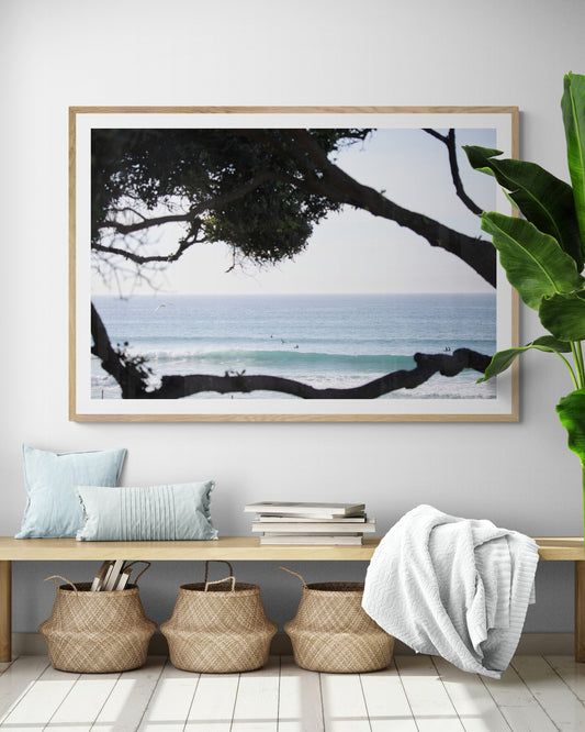 An ocean view reveals surfers on gentle waves, with a white bird soaring. Tree branches provide a natural, rustic foreground to the serene scene, taken at a beach in Del Mar, San Diego. Photograph in minimalist mock up.