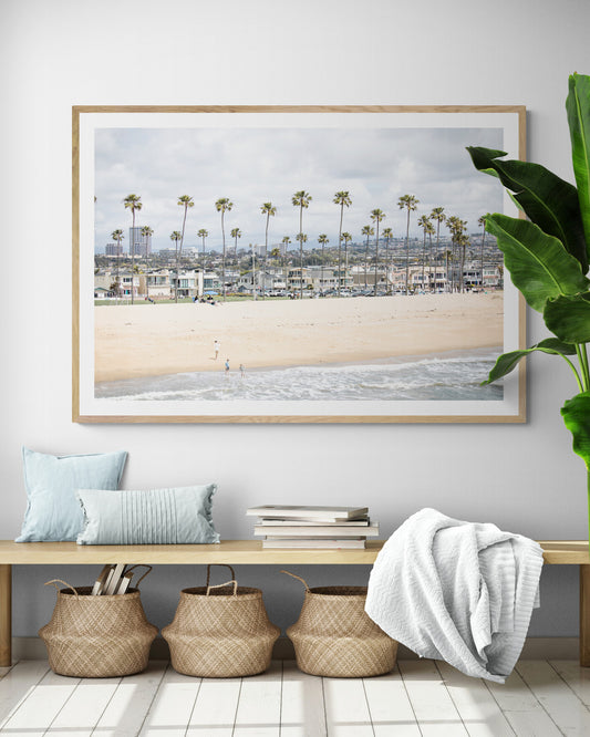 Landscape beach photography of Newport Beach, California, showing a wide sandy beach, a long line of tall palm trees, and coastal homes. Photograph in minimalist mock up.