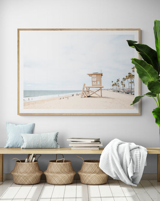 A wide landscape coastal photography view of Newport Beach in Southern California, featuring a row of tall palm trees, a lifeguard tower, and people enjoying the calm beach. Photograph in minimalist mock up.