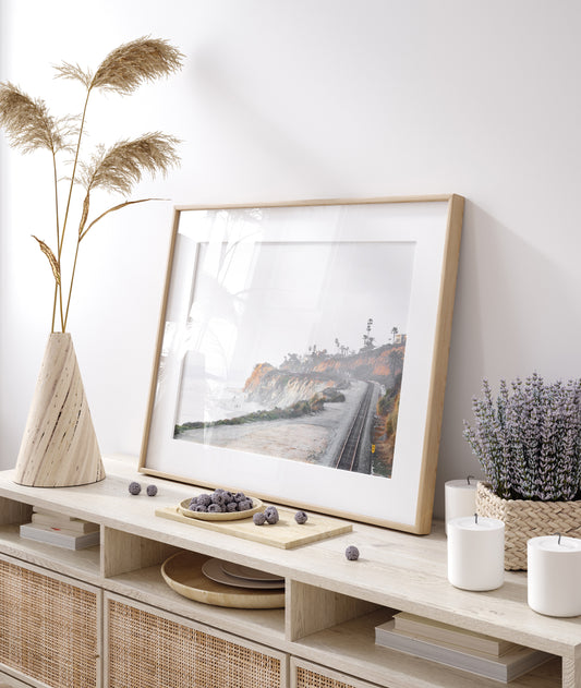 A slightly hazy, wide Landscape Coastal Photography shot shows a set of train tracks curving along a coastal cliffside, San Diego Coast. To the left, a sandy beach meets a light blue ocean waves with gentle waves, where a few small figures of people are visible. The cliffs are varied in color, with sections of tan, white, and hints of orange, topped with greenery and a few Palm Trees. The sky is a muted white, perfect for Minimalist Surf Wall Art. Photograph in minimalist mock up