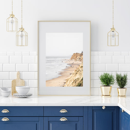 Fine art of Torrey Pines, San Diego California vertical print of hazy high-angle landscape beach photography captures a light blue ocean meeting a sandy beach with many beach goers. To the right, tall tan and brown cliffs rise as iconic coastal scenery. Photograph in minimalist framed kitchen mock up.