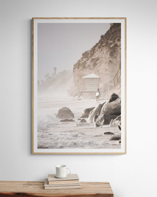 Vertical surf art print of a lone surfer carrying a surfboard, walking on a rocky, foggy shoreline at Moonlight State Beach with a lifeguard tower and cliff in the background. Photograph in minimalist mock up.