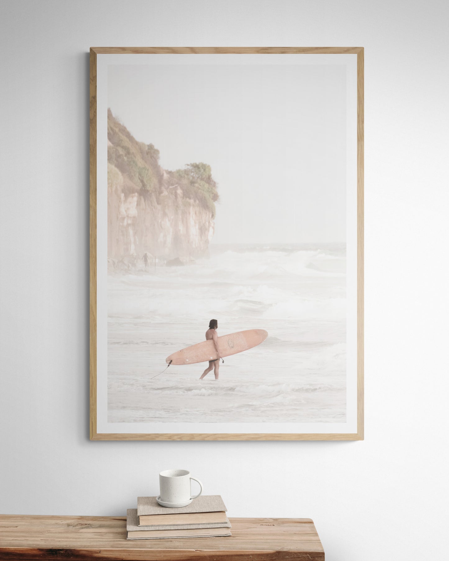 Vertical surf art print of a lone surfer carrying a surfboard, walking on a rocky, foggy shoreline at Moonlight State Beach with a lifeguard tower and cliff in the background. Photograph in minimalist mock up.