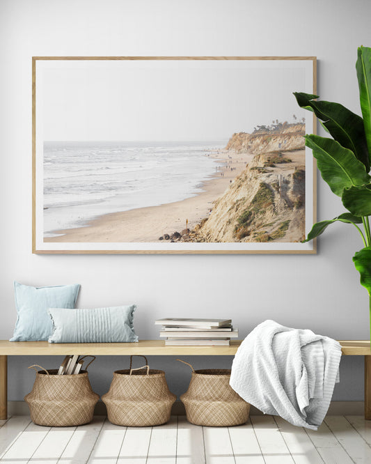 Fine art of Torrey Pines, San Diego California horizontal print of hazy high-angle landscape beach photography captures a light blue ocean meeting a sandy beach with many beach goers. To the right, tall tan and brown cliffs rise as iconic coastal scenery. Photograph in minimalist framed mock up.