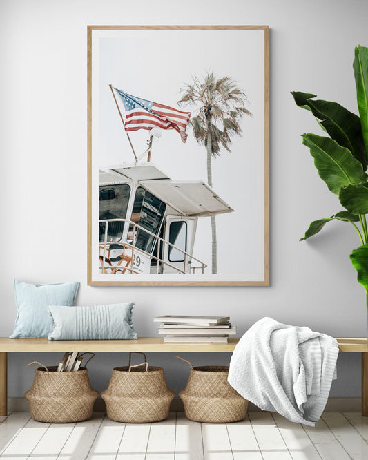 An American flag attached to a lifeguard tower with palm trees in the background, captured in California with a clear sky in the top half of the image. Photograph in minimalist mock up.