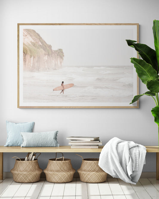 Horizontal surf art print of a lone surfer carrying a surfboard, walking on a rocky, foggy shoreline at Moonlight State Beach with a cliff in the background. Photograph in minimalist mock up.