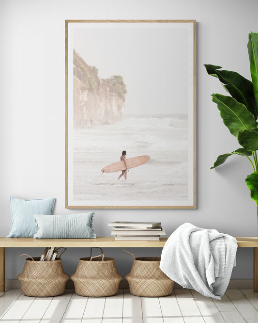 Vertical surf art print of a lone surfer carrying a surfboard, walking on a rocky, foggy shoreline at Moonlight State Beach with a lifeguard tower and cliff in the background. Photograph in minimalist mock up.