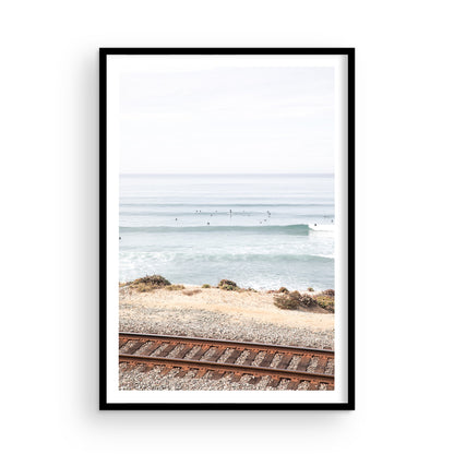 A framed print of a beach scene with waves and surfers, displayed on a train track in Del Mar, San Diego. Photograph in minimalist mock up.