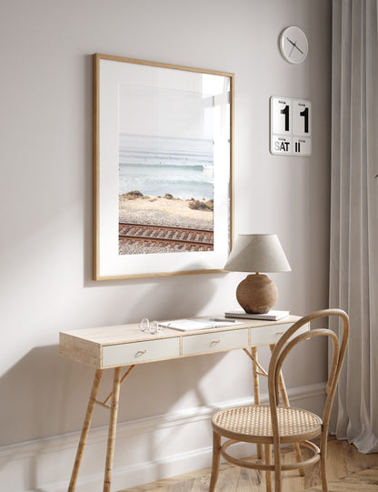 A framed print of a beach scene with waves and surfers, displayed on a train track in Del Mar, San Diego. Photograph in minimalist mock up.