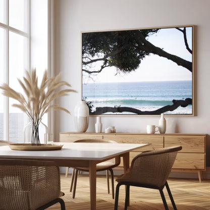 An ocean view reveals surfers on gentle waves, with a white bird soaring. Tree branches provide a natural, rustic foreground to the serene scene, taken at a beach in Del Mar, San Diego. Photograph in minimalist mock up.