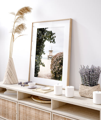 Vertical coastal photography of a Newport Beach lifeguard tower on the sand, framed by lush green and flowering foliage. Photograph in minimalist mock up.